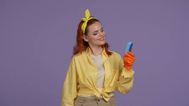 Everyday cleaning and housekeeping concept. Woman in casual clothing and rubber gloves holding blue sponge, looking at it smiling. Isolated on purple background. - Foto, afbeelding