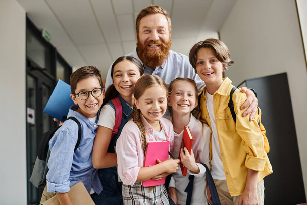 A man teacher engages with a diverse group of children in a lively hallway, imparting knowledge and guidance in a bright educational environment. - Photo, Image