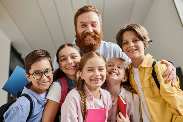 Μια ποικιλόμορφη ομάδα παιδιών στέκεται δίπλα σε έναν άνδρα σε ένα φωτεινό περιβάλλον στην τάξη - Φωτογραφία, εικόνα