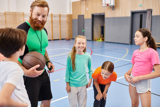 Nauczyciel trzyma koszykówkę, podczas gdy zróżnicowana grupa dzieci stoi wokół niego w jasnej, żywej sali lekcyjnej.. - Zdjęcie, obraz