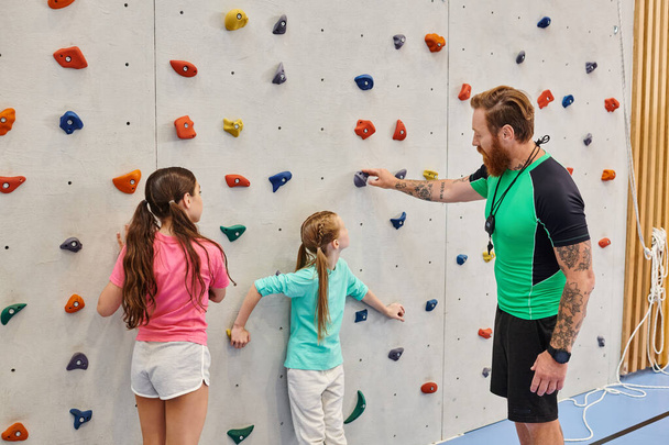 A man teacher instructs two young girls in front of a climbing wall in a bright, lively classroom setting. - Photo, Image