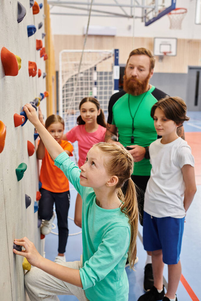 A man teacher instructs a diverse group of kids and adults as they stand around a climbing wall, preparing to embark on an adventurous climbing challenge. Bright, lively classroom setting adds to the excitement. - Photo, Image