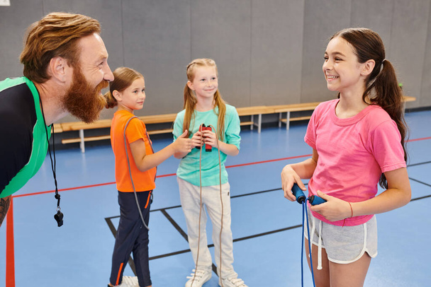 Een bebaarde man geeft enthousiast les aan een groep verschillende kinderen in een levendige gymzaal en weet hun aandacht te trekken met boeiende lessen.. - Foto, afbeelding