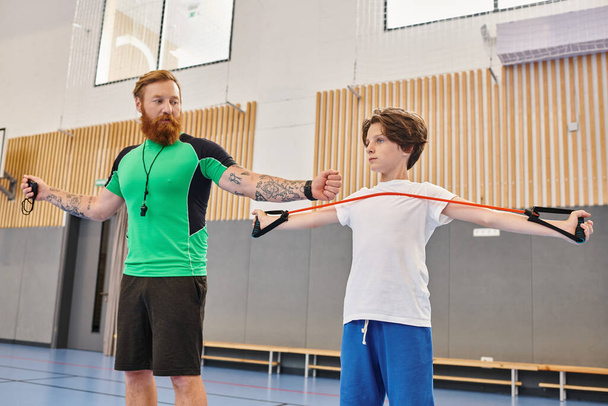 A bearded instructor with tattoos guides a young student through arm stretches in a vibrant school gymnasium. - Photo, Image