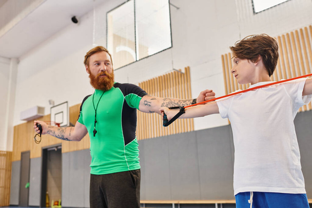 Un instructeur barbu donnant des conseils à un enfant concentré pratiquant dans une salle de gym ensoleillée. - Photo, image