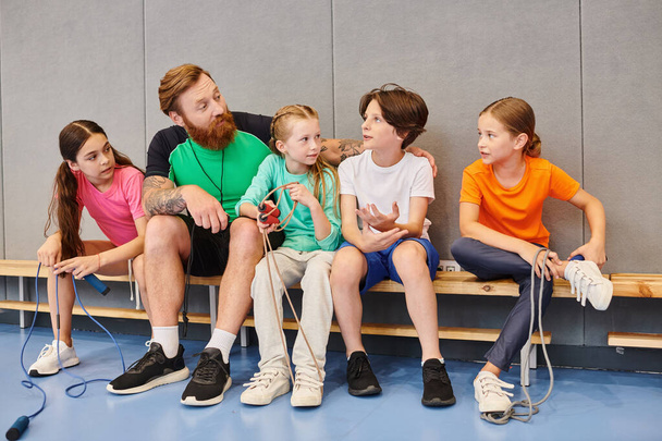 A diverse group of children sitting attentively on a bench, listening to their male teacher in a bright, lively classroom setting. - Photo, Image