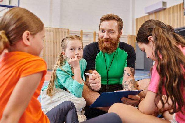 Egy szakállas férfi ül a kosárlabdapályán, körülvéve változatos gyerekekkel, mentorálva és edzve őket energikus és pozitív légkörben.. - Fotó, kép