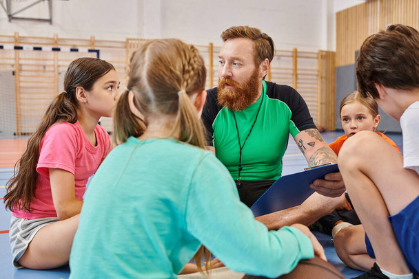 Een gevarieerde groep mensen, waaronder kinderen en hun mannelijke leraar, zitten om elkaar heen in een fitnessruimte, bezig met een levendige en levendige leersessie. - Foto, afbeelding