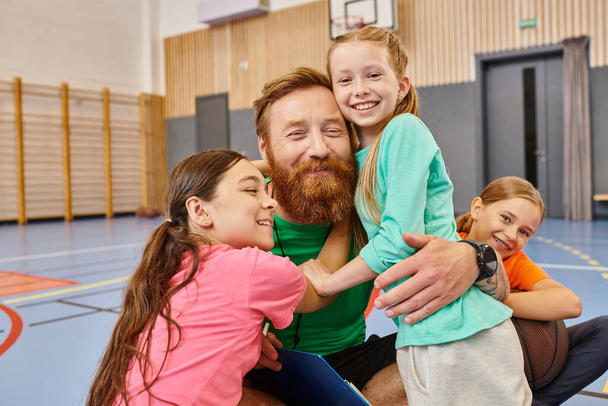 Un insegnante barbuto a braccia aperte, che abbraccia calorosamente un gruppo eterogeneo di bambini in una vibrante aula piena di risate e gioia. - Foto, immagini