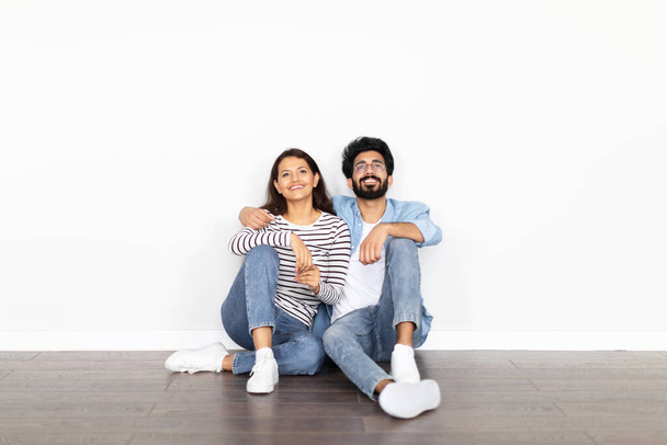 Happy positive beautiful loving young indian couple buying new house, sitting on floor in empty room in apartment, looking at copy space above them on white wall. Mortgage for millennials, rent - Photo, Image