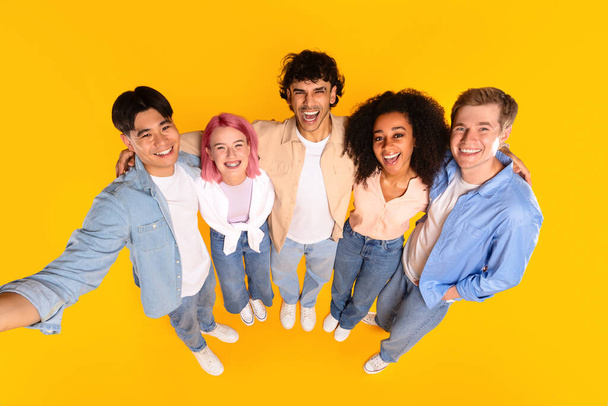 Boldog nemzetközi csoport tinédzser barátok ölelkeznek és szelfit készítenek, sárga stúdió háttér felett pózolnak, szem előtt tartva. Sokszínűség és barátság - Fotó, kép