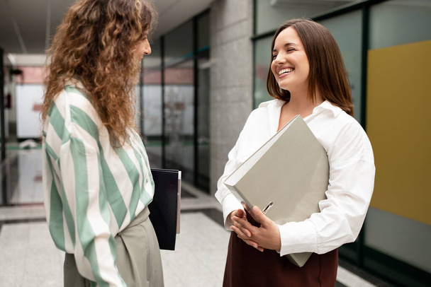 Business Communication. Two Happy European Businesswomen Talking And Laughing During Break At Office Coworking Interior, Standing And Holding Folders. Successful Career. Selective Focus - 写真・画像