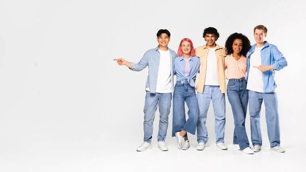 Look there. Group of excited multiracial friends pointing fingers aside at copy space and smiling at camera, standing on white background in studio, panorama - Foto, imagen