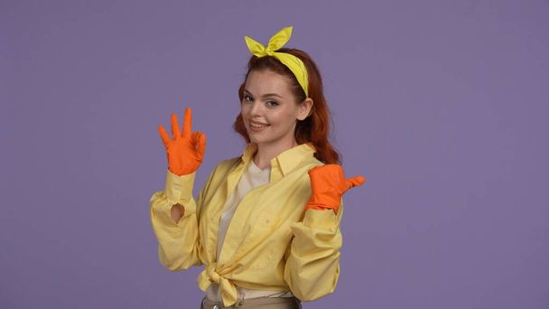 Everyday cleaning and housekeeping concept. Woman in casual clothing and rubber gloves showing ok sign pointing at the empty area, happy face. Isolated on purple background. - Foto, Imagen