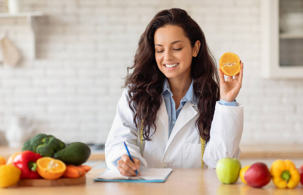 Εύθυμη Ευρωπαία ιατρός διατροφολόγος σε λευκό παλτό γράφοντας σχέδιο διατροφής, συνταγή με βιολογικά φρούτα, κρατώντας πορτοκάλι. Υγεία, απώλεια βάρους και επαγγελματική σύσταση - Φωτογραφία, εικόνα