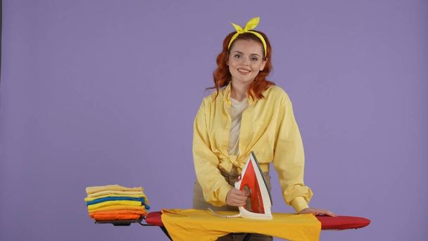 Everyday cleaning and housekeeping concept. Woman in casual clothing and headband standing and posing with iron and board, stack of colorful clothes next to her. Isolated on purple background. - Foto, Bild