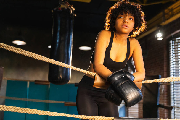 Savaşçı kadın gücü. Boks eldivenli Afrika kökenli Amerikalı kadın boksör boks ringinde iplere yaslanmış bekliyor ve dinleniyor. Güçlü ve güçlü bir kız. Vücut çalıştırma eğitimine uygun güç - Fotoğraf, Görsel