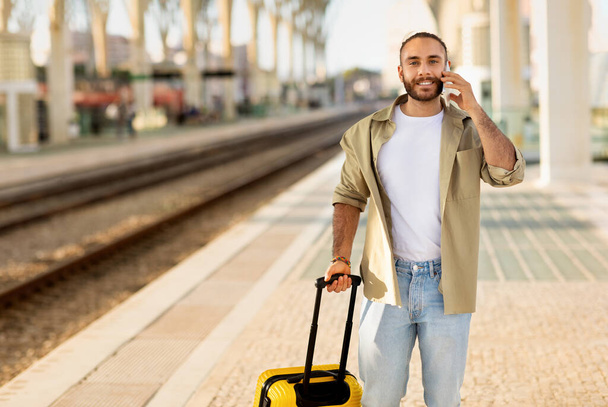 Glimlachende jonge blanke man met koffer gesprekken per telefoon, praten, wachten op vervoer op het station. Reisstijl, toestel voor reis, communicatie, goed nieuws, zakelijk - Foto, afbeelding