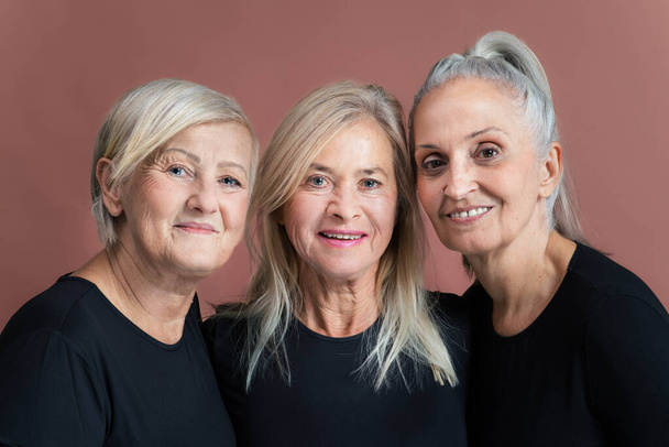 Studio portrait of three beautiful senior women with gray hairs in black tops. Elderly female friends holding each other looking at camera. Isolated on a brown pink background. Copy space. - 写真・画像