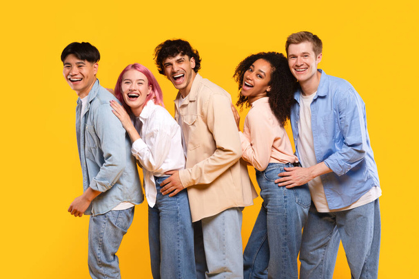 Group of joyful multiracial teen friends standing together over yellow background, posing and laughing, having fun and fooling in studio, looking at camera - Fotoğraf, Görsel