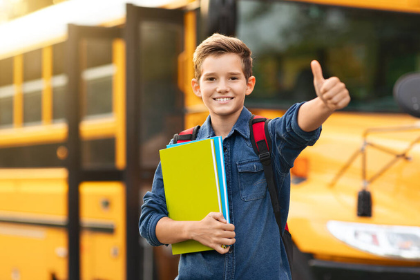 Sonriente niño preadolescente de pie cerca de autobús escolar amarillo y mostrando el pulgar hacia arriba en la cámara, colegial feliz celebración de libros de trabajo en las manos, Disfrutar de estudio, Recomendar programas educativos, Copiar el espacio - Foto, imagen