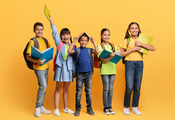 Fröhlich lächelnde multirassische Schulkinder mit Rucksäcken und Büchern posieren isoliert auf gelbem Studiohintergrund. Verschiedene Kinder im Vorschulalter gehen gemeinsam zur Schule. Bildung für Kinder - Foto, Bild