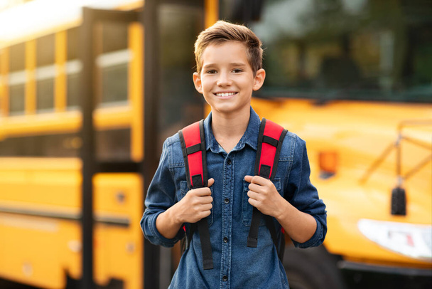 Menino pré-adolescente sorridente feliz em pé perto de ônibus escolar amarelo ao ar livre, criança masculina alegre com mochila posando fora enquanto vai para as aulas, olhando para a câmera, estudante desfrutando de estudo, espaço de cópia - Foto, Imagem