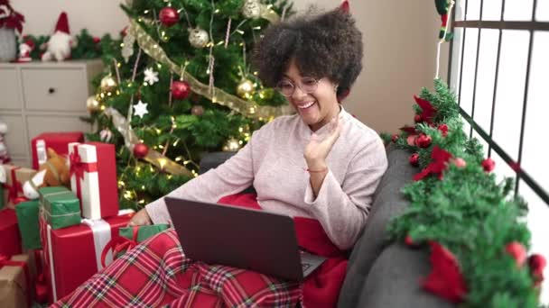 Young african american woman having video call holding gift sitting by christmas tree at home - Footage, Video