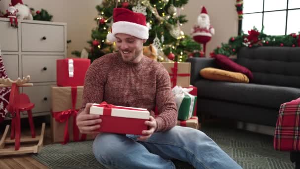 Nuori valkoihoinen mies purkaa lahja istuu joulukuusi kotona - Materiaali, video