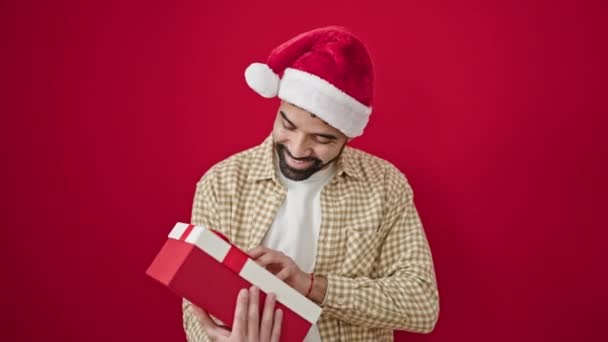 Młody Hiszpan uśmiecha się pewnie nosząc świąteczny kapelusz słysząc dźwięk prezentu nad odizolowanym czerwonym tłem - Materiał filmowy, wideo