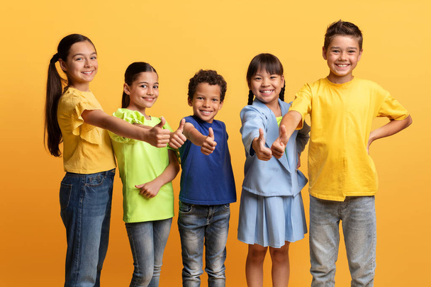 Большое дело, волнующее предложение для детей. Счастливые многонациональные дети, мальчики и девочки в повседневных нарядах, показывающие большой палец вверх и улыбающиеся в камеру, изолированные на желтом фоне - Фото, изображение