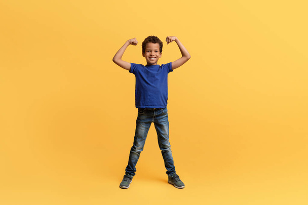 Strong confident cute athletic african american school aged boy raising hands up, preteen kid showing his biceps and smiling at camera, isolated on yellow background. Sports lifestyle, dreams - Photo, Image