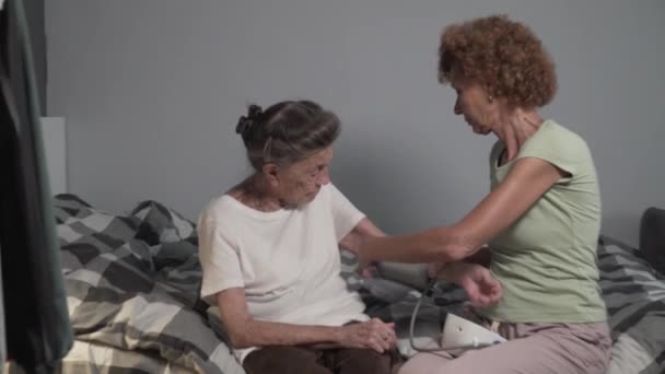 La mujer madura ayuda a la mujer mayor a medir la presión arterial mediante el esfigmomanómetro de electrodomésticos médicos. La hija mayor está cuidando a la vieja madre enferma. Enfermería examen médico enfermedad hipertensión. - Imágenes, Vídeo