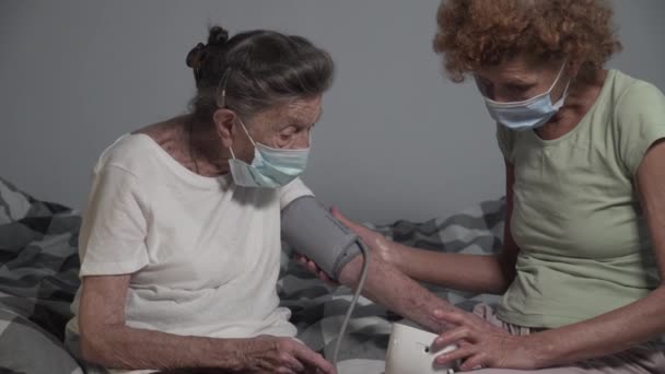 Зрелая женщина-сиделка в медицинской маске проверяет кровяное давление пожилых пациентов во время пандемии, сидит на кровати в доме престарелых. Старшую женщину посетил специалист по уходу на дому. Старые люди с гипертонией. - Кадры, видео