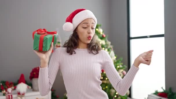 Young beautiful hispanic woman dancing by christmas tree holding gift at home - Footage, Video