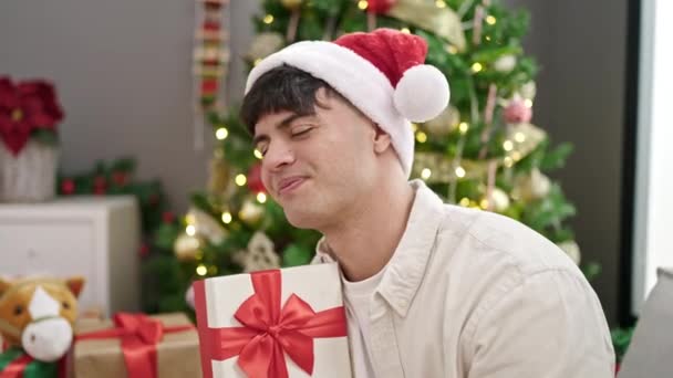 Νεαρός Ισπανός άνδρας χαμογελά σίγουρος κρατώντας χριστουγεννιάτικο δώρο στο σπίτι - Πλάνα, βίντεο