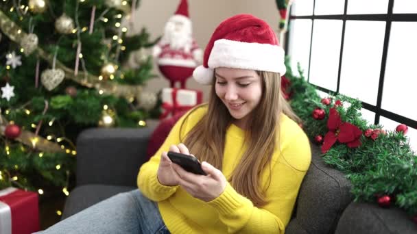 Nuori blondi nainen käyttää älypuhelinta istuu sohvalla joulupuulla kotona - Materiaali, video