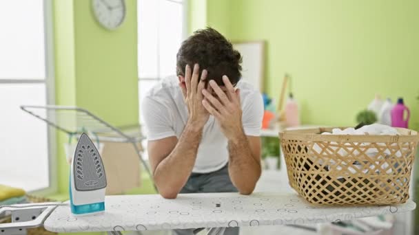 Joven hispano apoyado en tabla de planchar estresado en la lavandería - Imágenes, Vídeo
