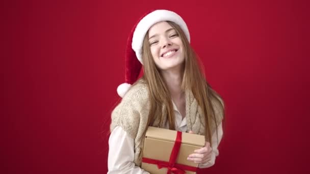 Mladá blondýna žena na sobě vánoční klobouk drží dárek přes izolované červené pozadí - Záběry, video