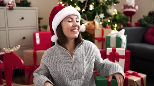 Joven mujer hispana hermosa sonriendo confiada sentada en el suelo por el árbol de Navidad en casa - Metraje, vídeo