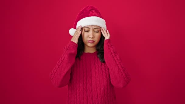 Fiatal gyönyörű spanyol nő visel karácsonyi kalap, amelynek fejfájás felett elszigetelt piros háttér - Felvétel, videó