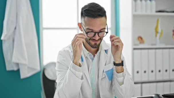 Jonge Spaanse man dokter zittend op tafel met armen gekruist gebaar het nemen van een bril af in de kliniek - Video