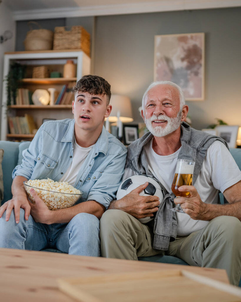 Καυκάσιος έφηβος και ηλικιωμένος παππούς και εγγονός κάθονται στον καναπέ-κρεβάτι στο σπίτι να παρακολουθήσουν ποδόσφαιρο παιχνίδι κατέχουν ποπ κορν μπολ και ποτήρι μπύρα διασκεδάζουν οικογενειακό δέσιμο άνδρες - Φωτογραφία, εικόνα