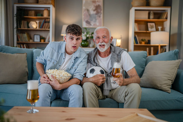Кавказский подросток и старший мужчина дедушка и внук сидеть на диване кровати дома смотреть футбол провести попкорн миску и бокал пива весело семейные связи мужчин - Фото, изображение