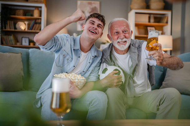 Kaukasischer Teenager und älterer Mann Großvater und Enkel sitzen zu Hause auf dem Schlafsofa und beobachten Fußballspiel halten Popcornschale und Glas Bier haben Spaß Familie Bindung Männer - Foto, Bild