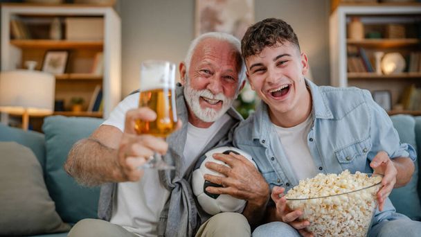 Caucasian teenager and senior man grandfather and grandson sit on sofa bed at home watch football soccer game hold popcorn bowl and glass of beer have fun family bonding males men - Foto, afbeelding