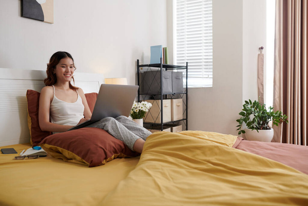 Giovane donna allegra seduta sul letto quando il video chiama amico o collega - Foto, immagini
