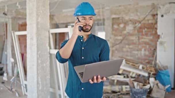 Joven arquitecto hispano usando casco usando laptop hablando por teléfono en el sitio de construcción - Imágenes, Vídeo