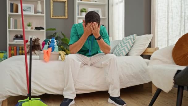 Junge hispanische Mann professionelle Reinigungskraft sitzt auf Bett gestresst im Schlafzimmer - Filmmaterial, Video