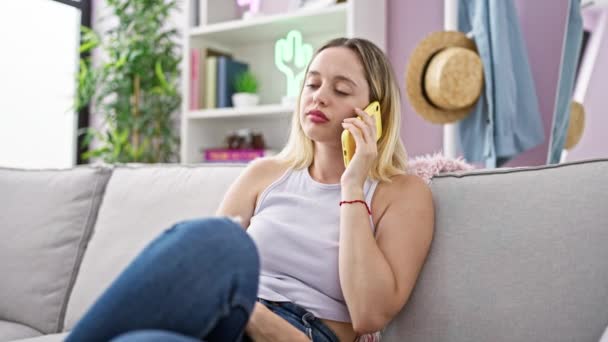 Νεαρή ξανθιά γυναίκα που μιλάει στο τηλέφωνο κάθεται στον καναπέ χασμουριέται στο σπίτι - Πλάνα, βίντεο
